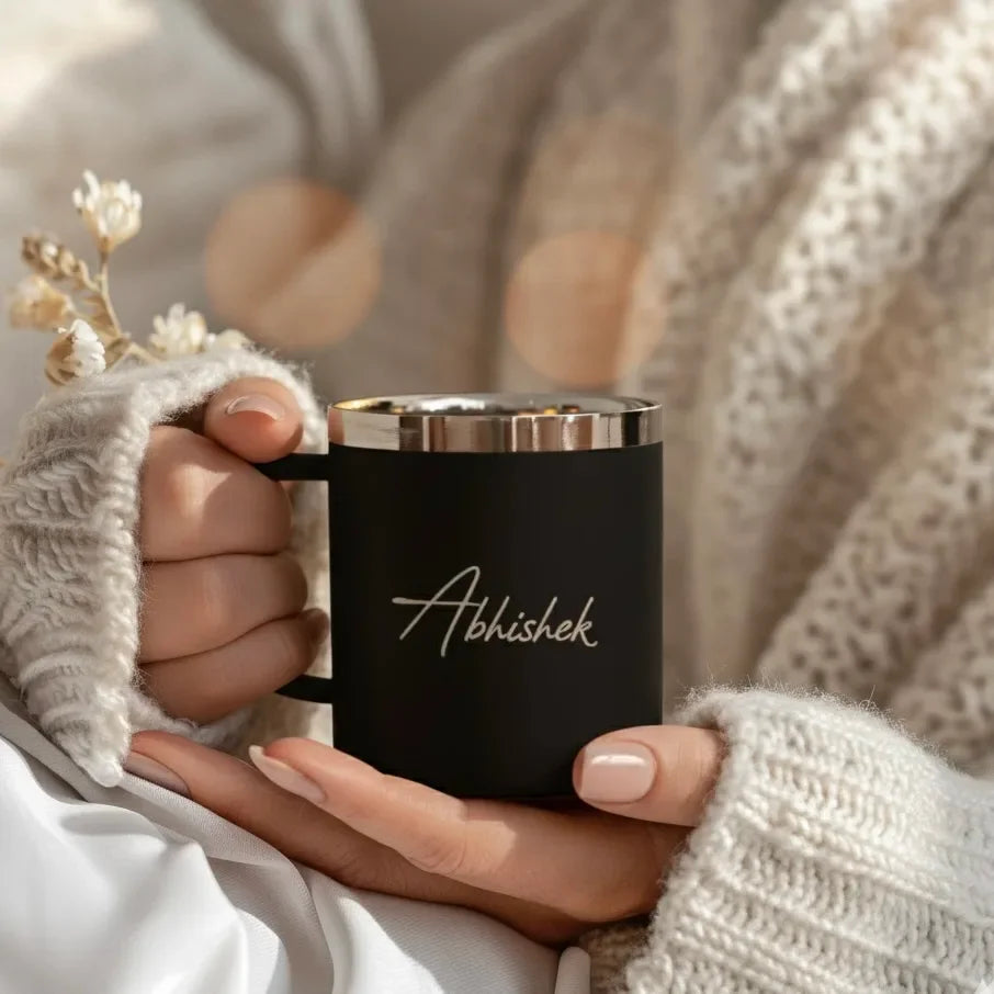 Person holding a black mug with 'Abhishek' written on it, wearing beige knitted sleeves.