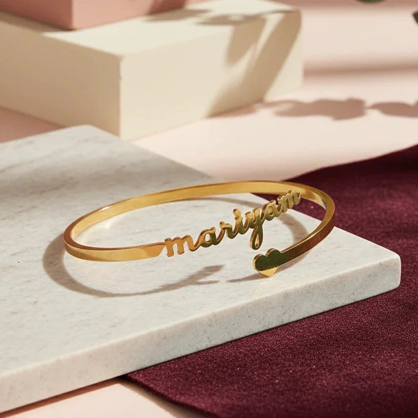 Gold bracelet with 'maryam' engraving on a marble surface with a pink and green leafy background