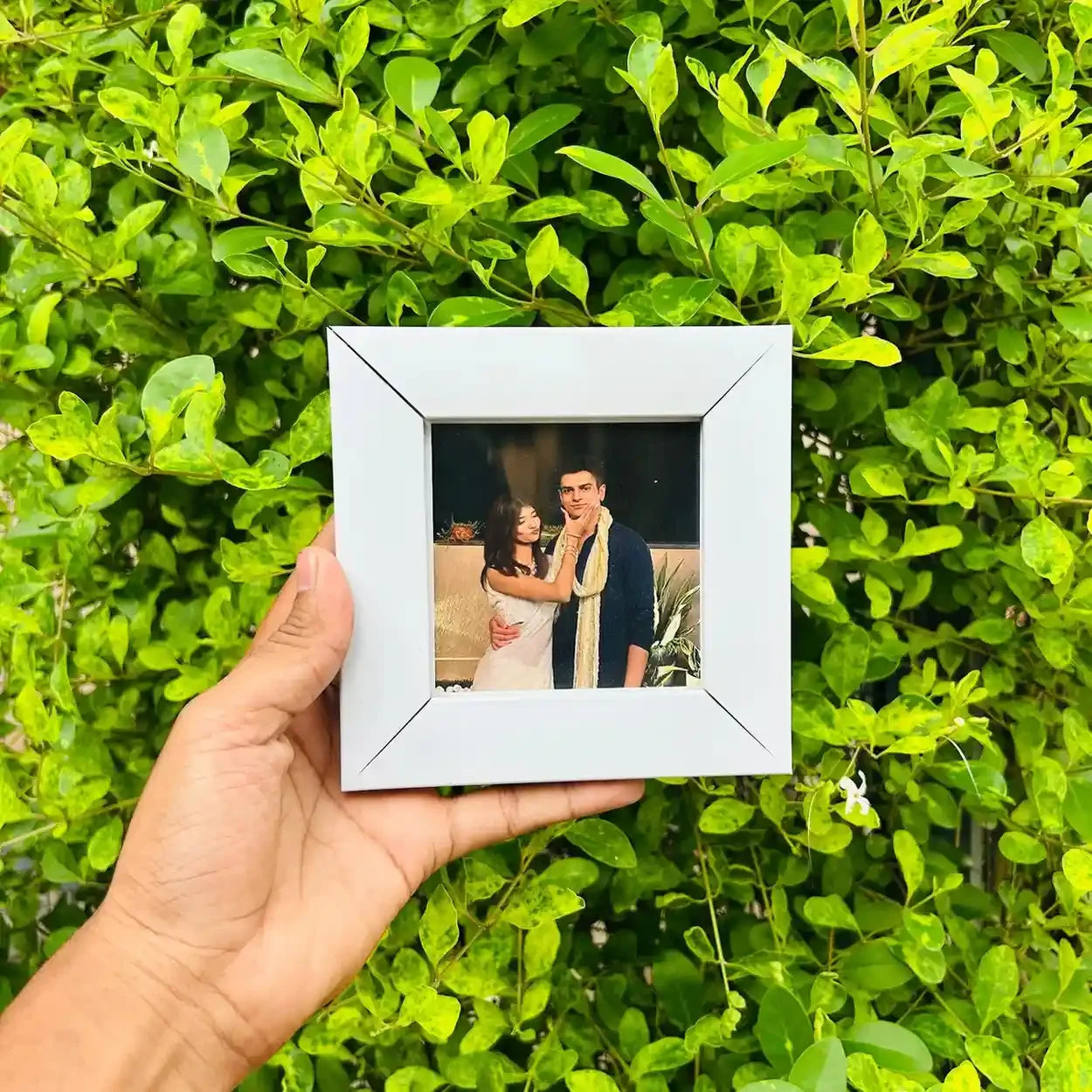 Hand holding a white photo frame with a picture of a couple against a green leafy background