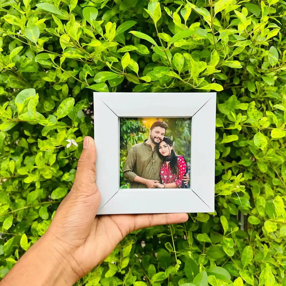 Hand holding a white photo frame with a couple's picture against a green leafy background