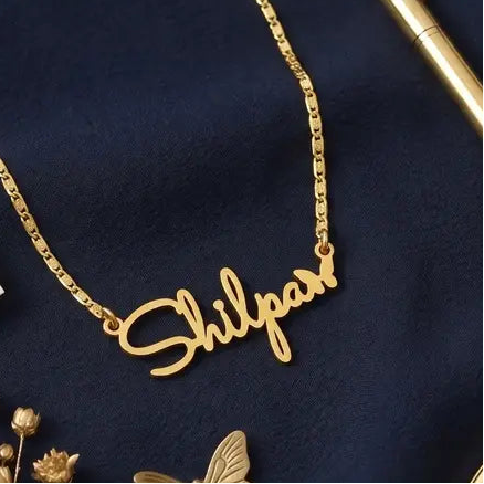 Gold necklace with 'Shilpan' tag, pen, and butterfly on dark blue fabric